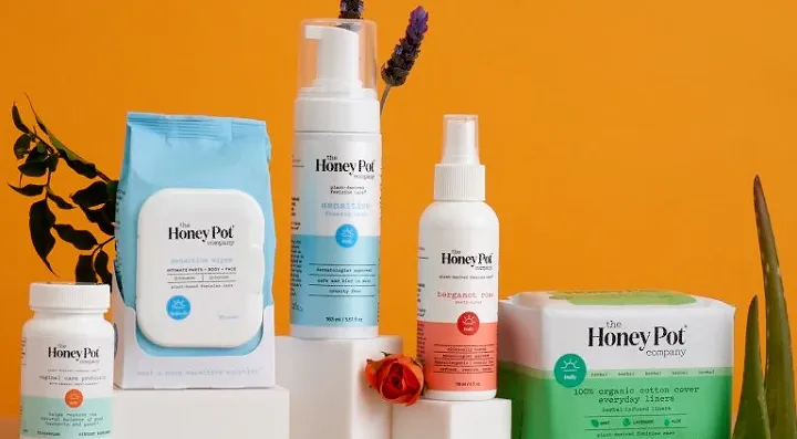Various Honey Pot company personal care products including wipes, foam, spray, and pads displayed against an orange background with some plant decor.